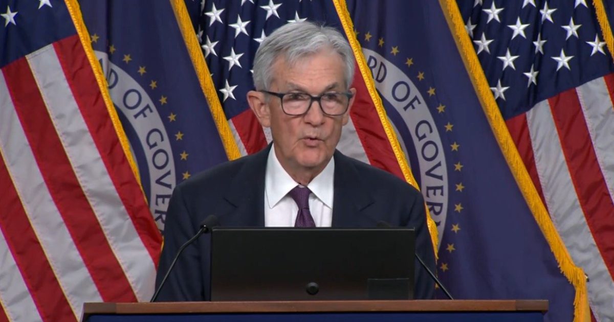 Federal Reserve Chairman delivering a speech at a press conference, with American flags and the Federal Reserve emblem in the background.