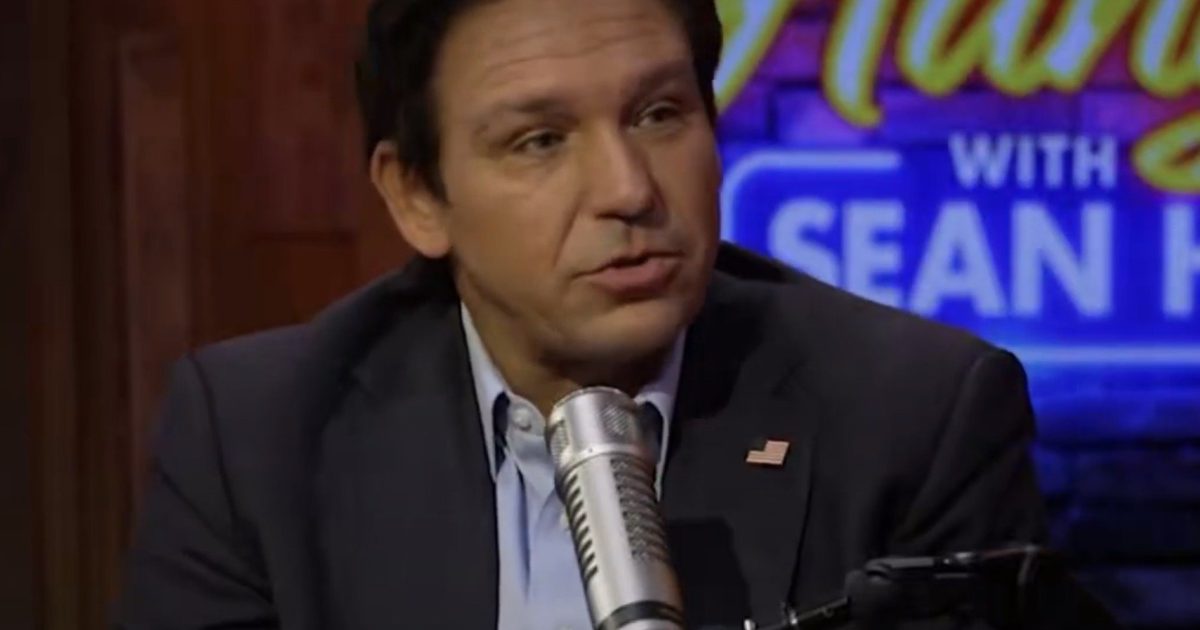Ron DeSantis speaking during an interview on a talk show, with a microphone in front of him and a colorful background.