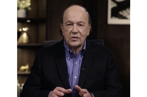 Man in a black blazer and blue shirt speaking confidently in an office setting, with decorative shelves in the background.