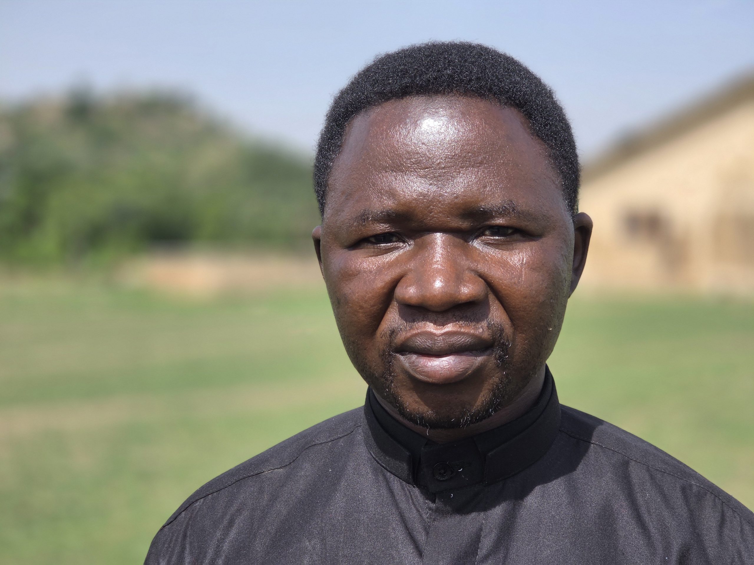 Portrait of a man wearing a black shirt standing outdoors against a blurred green background, highlighting his serious expression and natural setting.