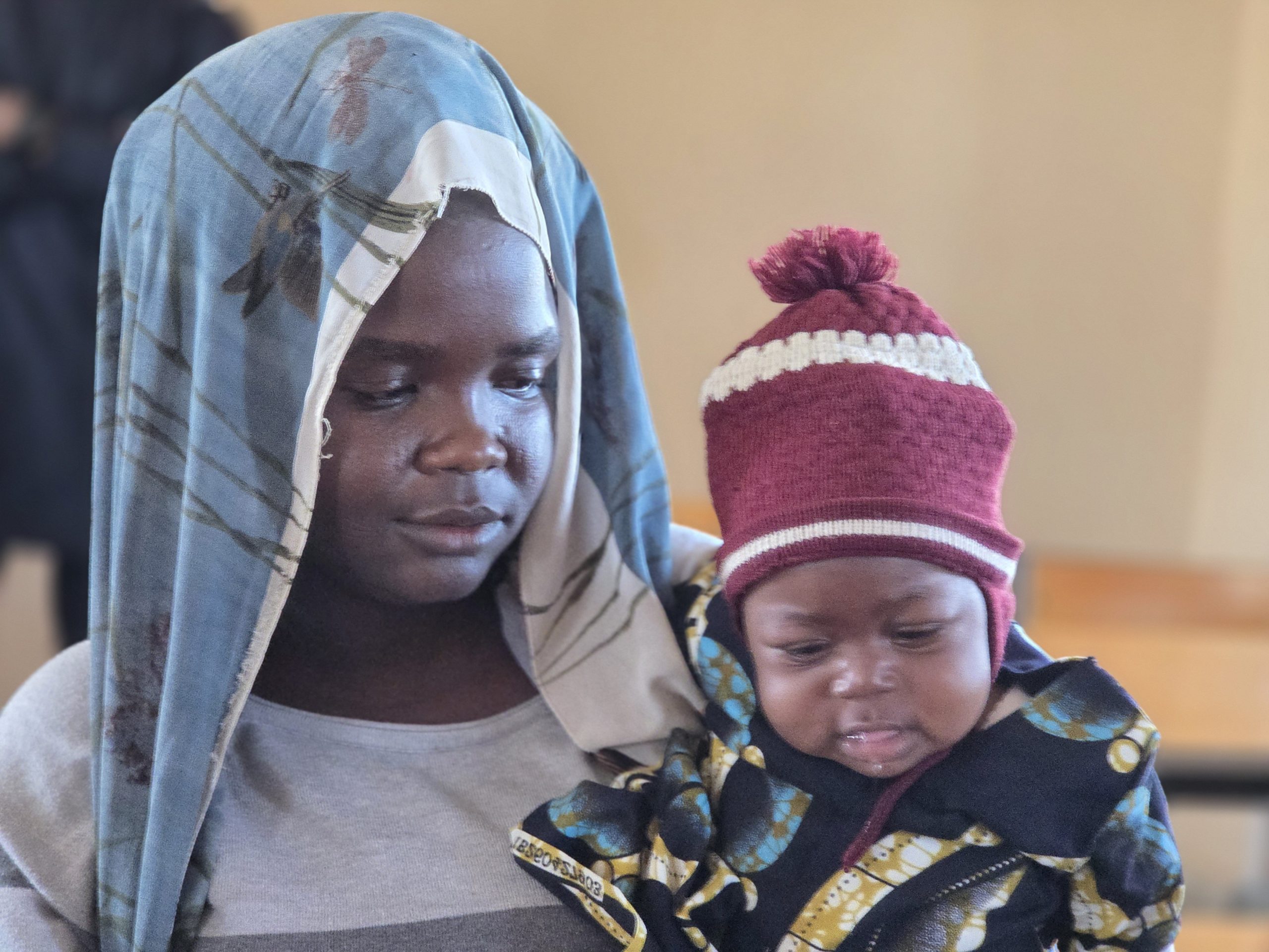 A mother wearing a blue scarf holds her baby, who is dressed in a colorful jacket and a red hat, in a warm indoor setting.