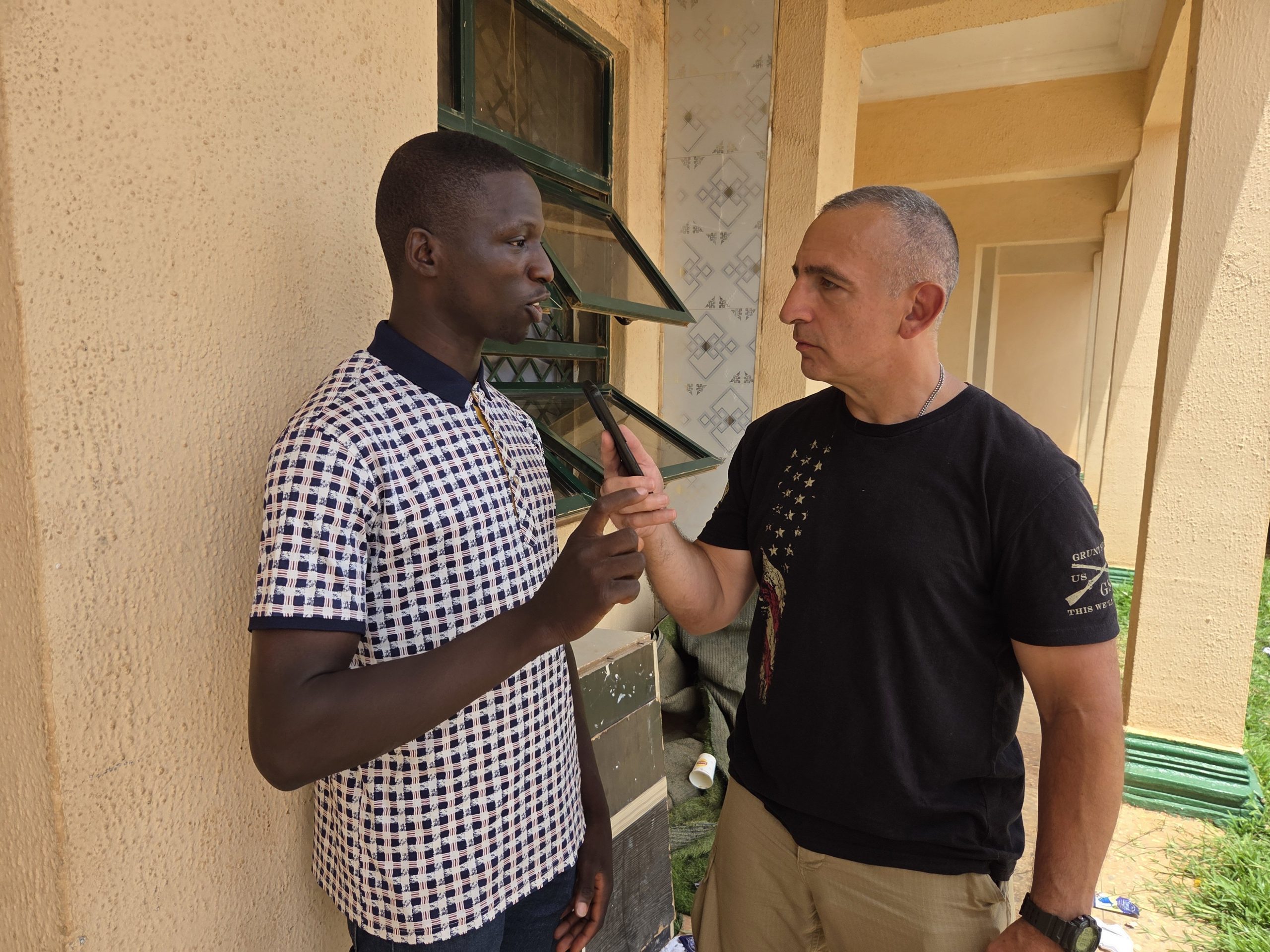 Two men engage in a serious conversation outside a building, with one gesturing while the other listens intently.