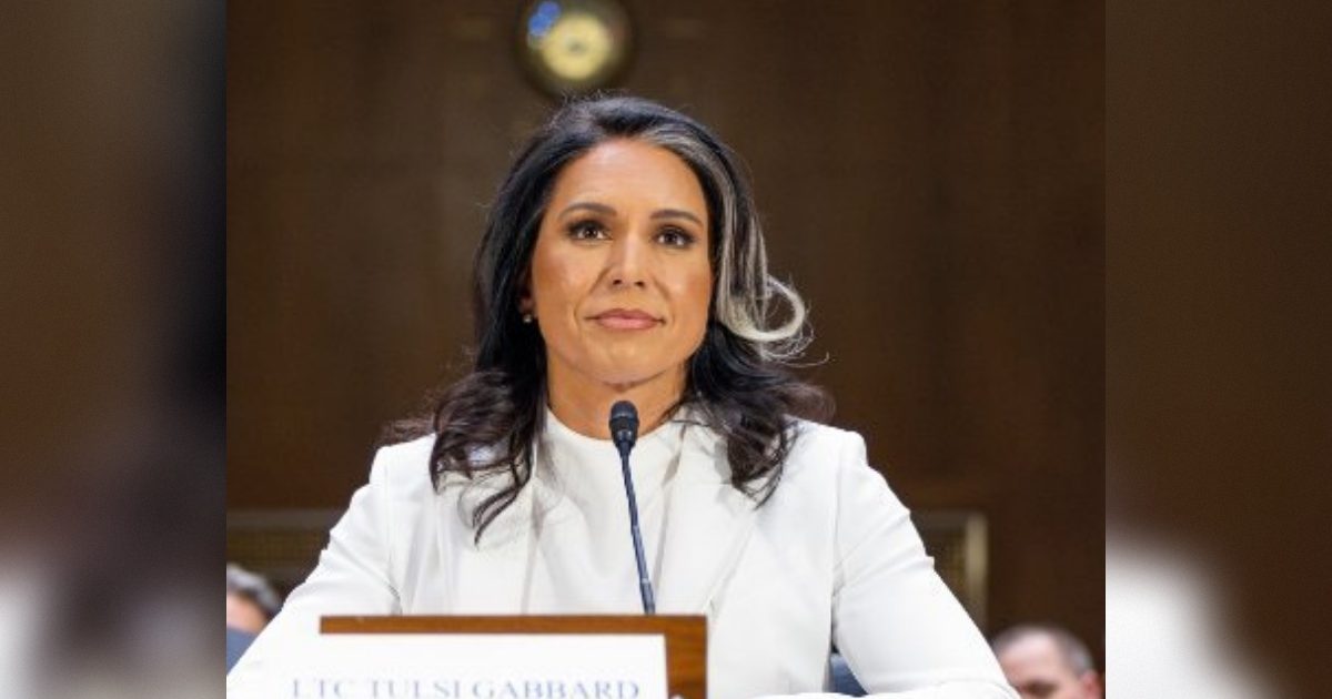 LTC Tulsi Gabbard testifies during a congressional hearing, seated at a table with a microphone, dressed in a white blazer, showcasing a serious expression.