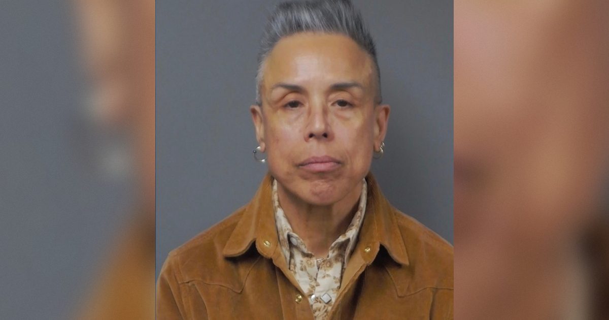 Mugshot of an individual with short gray hair, wearing a brown suede jacket and patterned shirt, against a plain background.