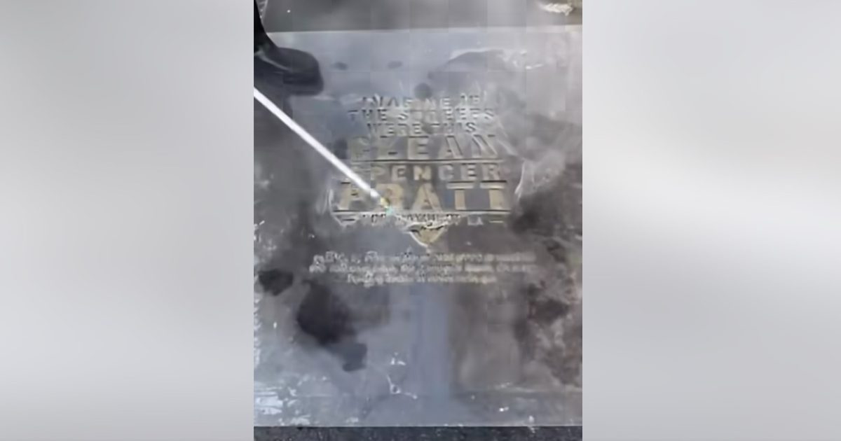A person cleaning a commemorative plaque for Dean Spencer Ball using a pressure washer, revealing the engraved details.