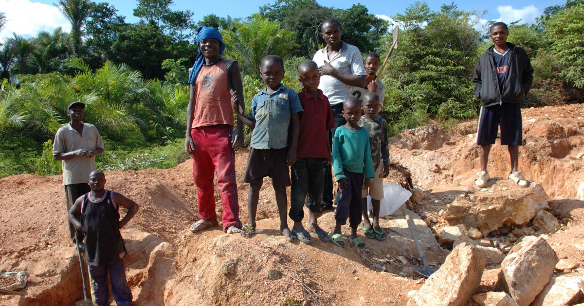 Group of children and adults standing on a mining site surrounded by rocks and vegetation, showcasing community labor and engagement in resource extraction activities.