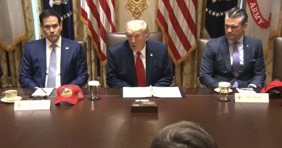Donald Trump and two officials seated at a table during a meeting, with flags in the background and a red cap visible on the table.