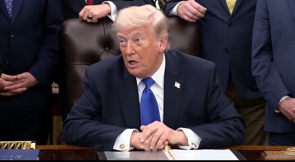 Donald Trump speaking during a press event at a desk surrounded by supporters, highlighting a significant announcement or policy discussion.