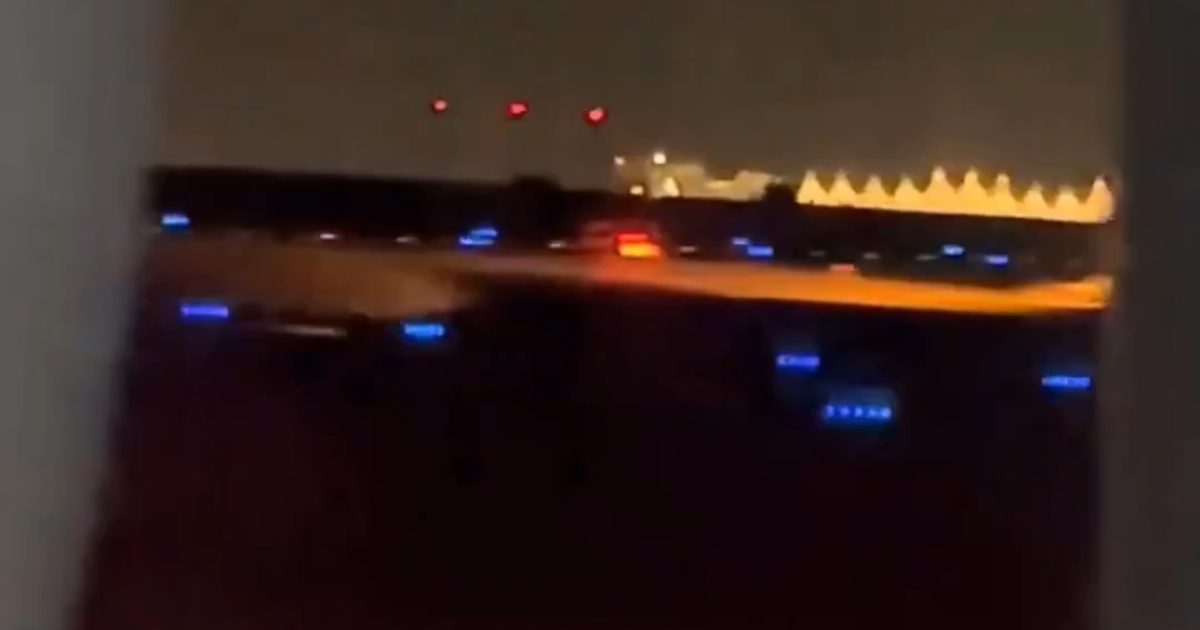 Nighttime airport runway view with illuminated taxiway lights and red signal lights in the background.
