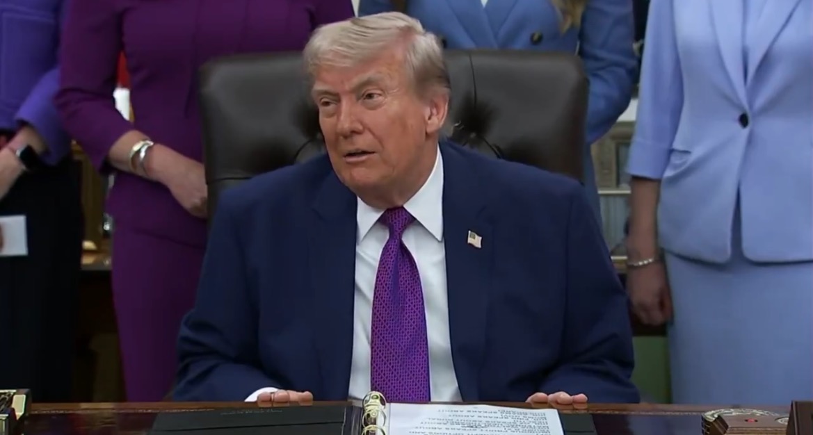 Donald Trump seated at a desk during an official event, discussing policies with advisors in formal attire.