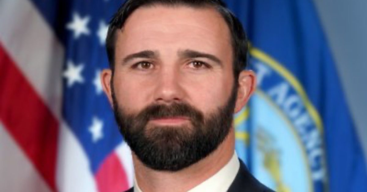 Professional portrait of a man with a beard in front of American and agency flags, representing leadership and authority.