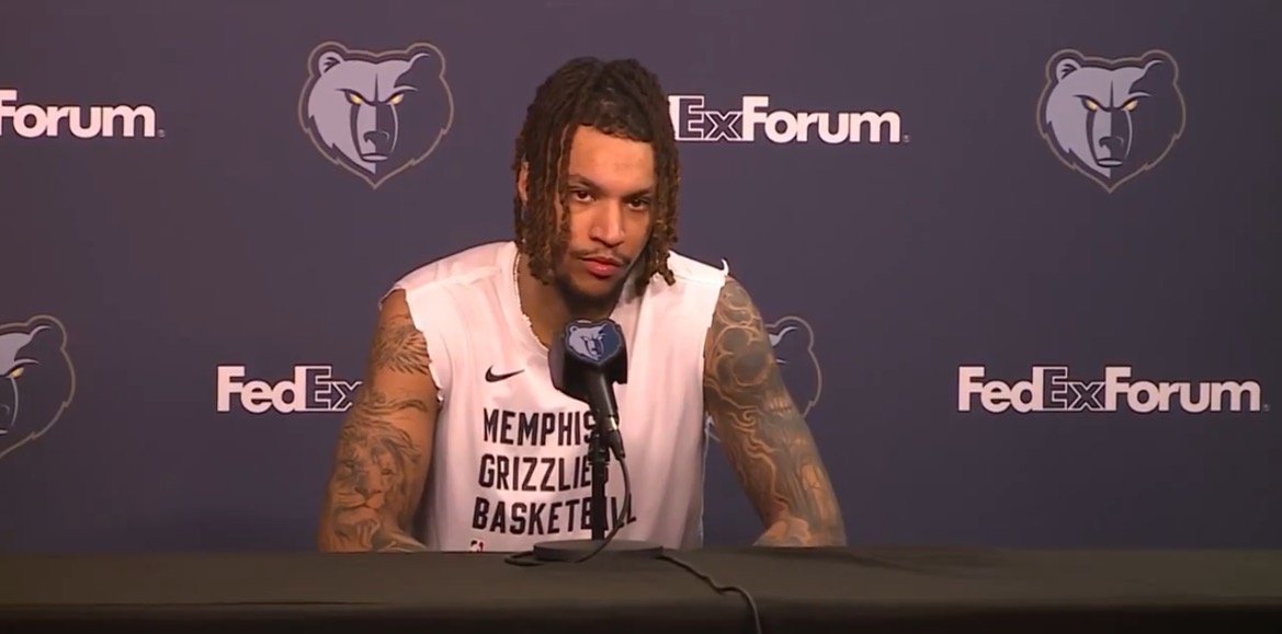 Memphis Grizzlies player at a press conference, discussing team updates and strategies with a backdrop featuring the team's logo and sponsor.