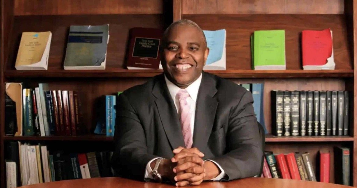 Smiling professional man in a suit seated at a desk with a bookshelf filled with various legal books in the background.