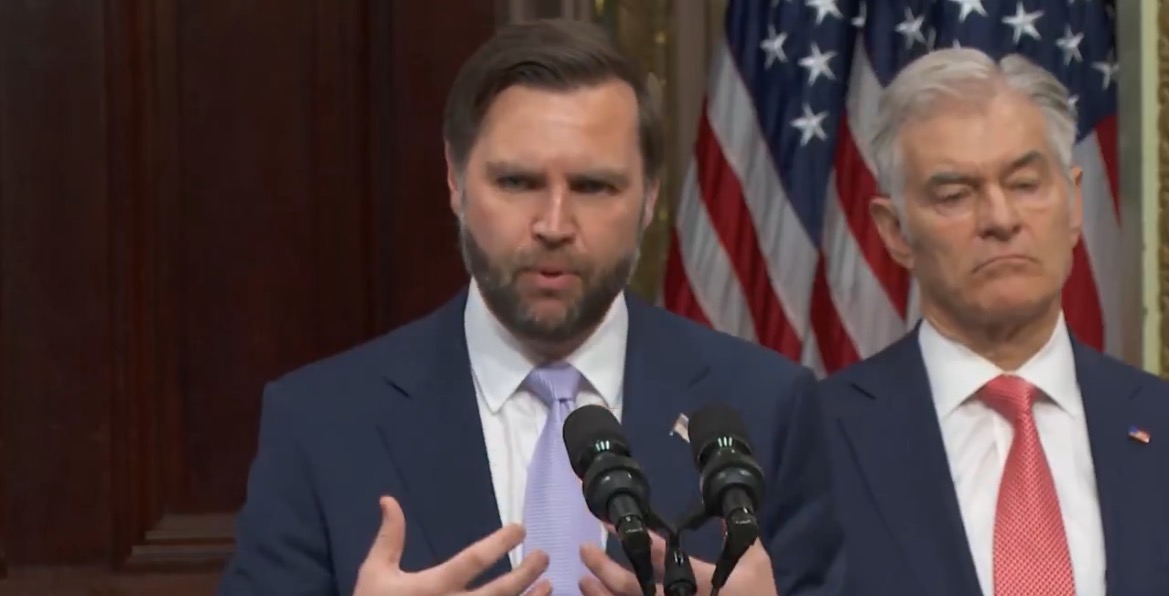 Speaker addressing an audience at a press conference, with American flags in the background, discussing economic policies alongside another official.