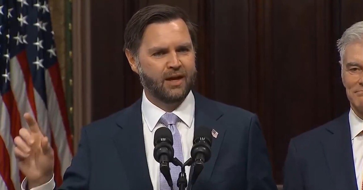 Man in a suit speaks at a podium with microphones, flags in the background, during a formal event or announcement.