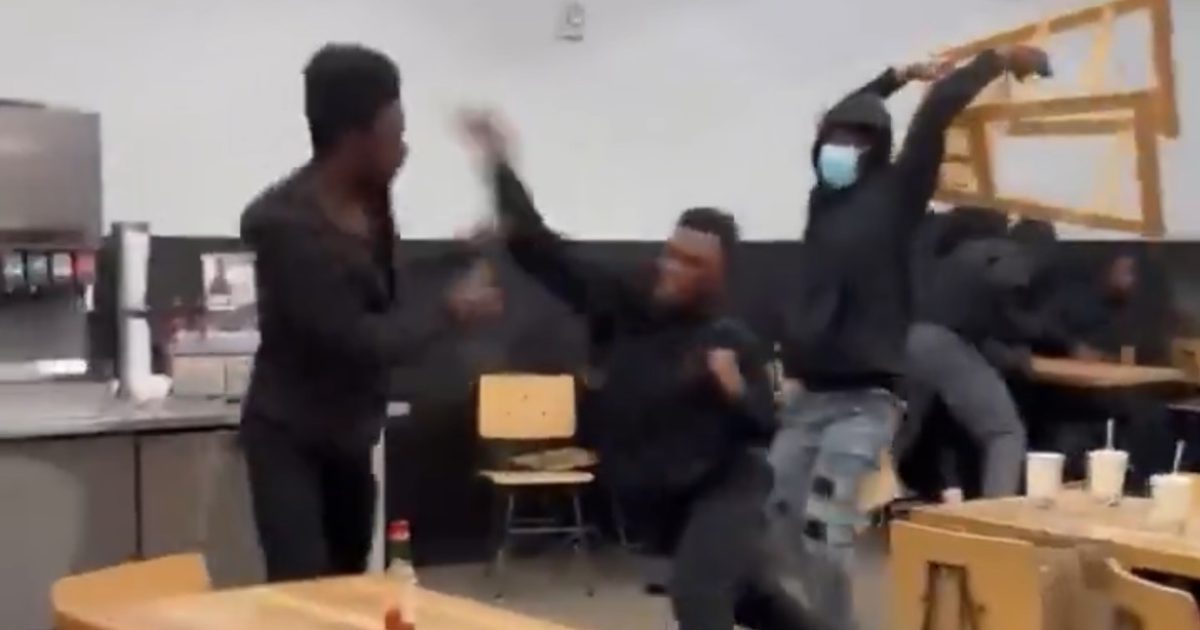A chaotic scene in a cafeteria with individuals in black clothing engaging in a physical altercation, chairs overturned and drinks scattered.
