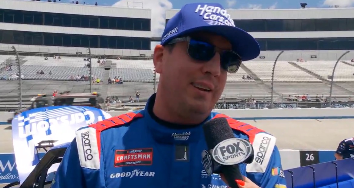 NASCAR driver being interviewed in front of a racetrack, wearing a blue racing suit and cap, with empty grandstands in the background.