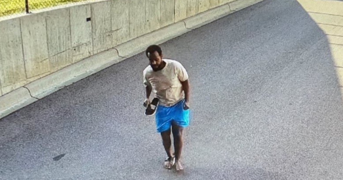 Man walking on a roadway wearing a beige t-shirt and blue shorts, holding a shoe in his left hand.