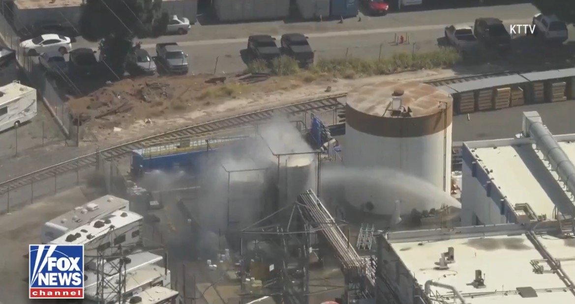 Industrial site with smoke rising from storage tanks, surrounded by parked vehicles and railway tracks in the background.