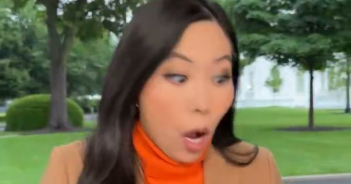 Surprised woman in a brown blazer and orange turtleneck, outdoors with trees in the background, expressing shock or amazement.
