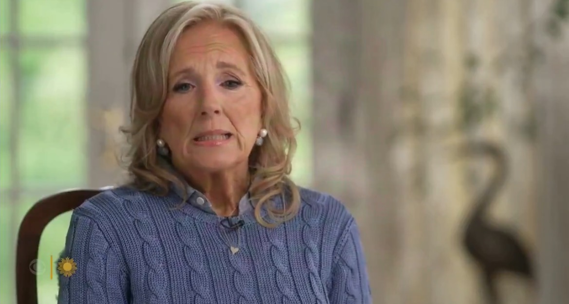 Woman with long blonde hair wearing a blue sweater, sitting indoors with a thoughtful expression, surrounded by natural light and soft decor.