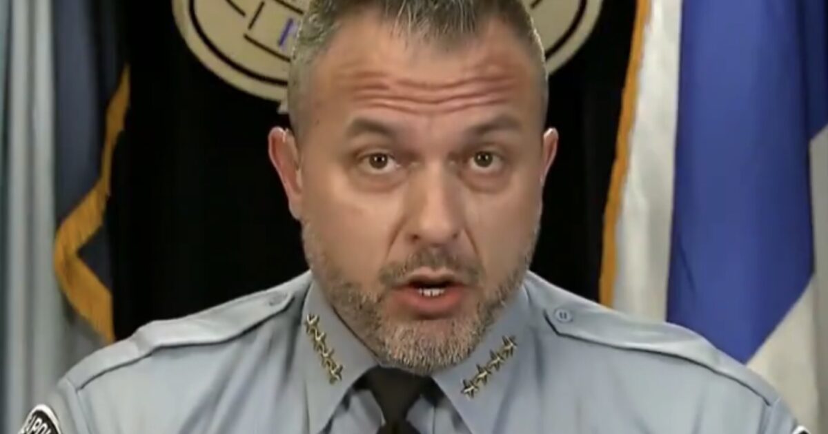 Police chief addressing the media, wearing a uniform with insignia, set against a backdrop of flags and an official seal.