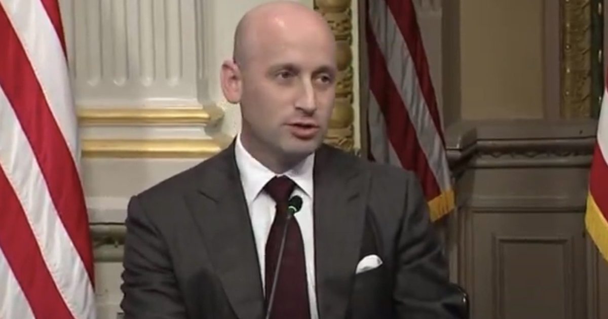Stephen Miller speaking at a press conference with American flags in the background, discussing policy issues.