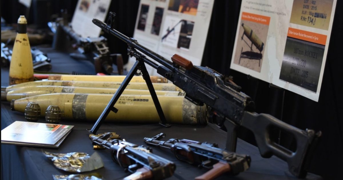 Display of various military weapons and ammunition, including rifles, grenades, and artillery shells, showcased on a table with informational posters in the background.
