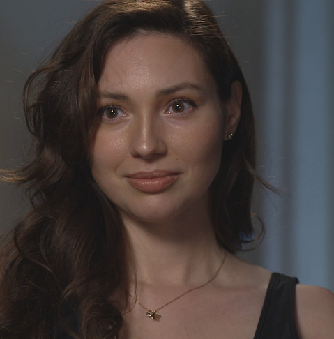 Portrait of a woman with long, wavy hair and a subtle smile, wearing a black top and a delicate necklace, set against a softly lit background.