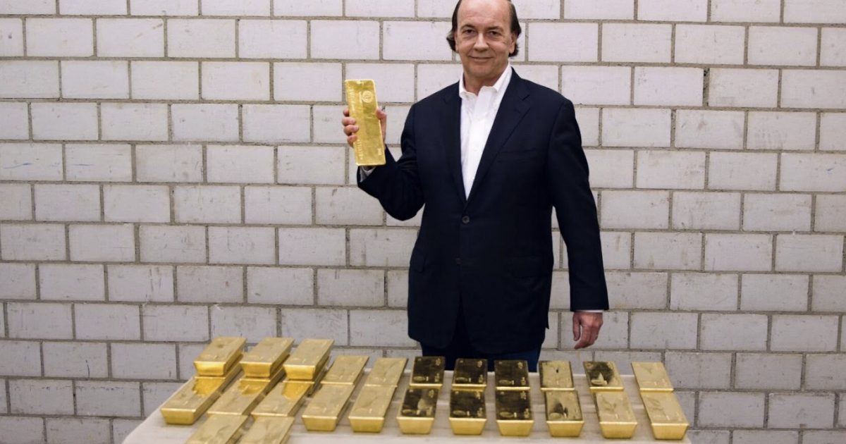 Man in a suit holding a gold bar stands in front of a table stacked with gold ingots against a brick wall backdrop.
