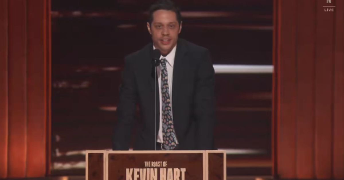 Comedian performing at the Roast of Kevin Hart, delivering jokes on stage with a microphone and a wooden podium in a well-lit setting.