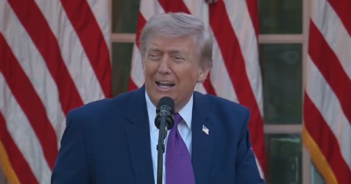 Donald Trump speaking at a podium with a microphone, surrounded by American flags in the background.