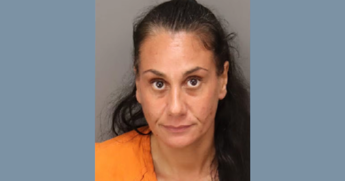 Mugshot of a woman with long dark hair, wearing an orange jumpsuit, looking directly at the camera against a neutral background.