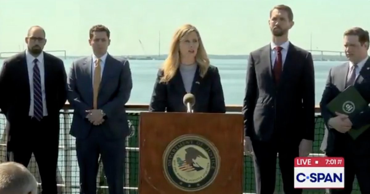 Press conference featuring a female speaker at a podium with the Department of Justice seal, surrounded by five male officials, set against a waterfront backdrop.