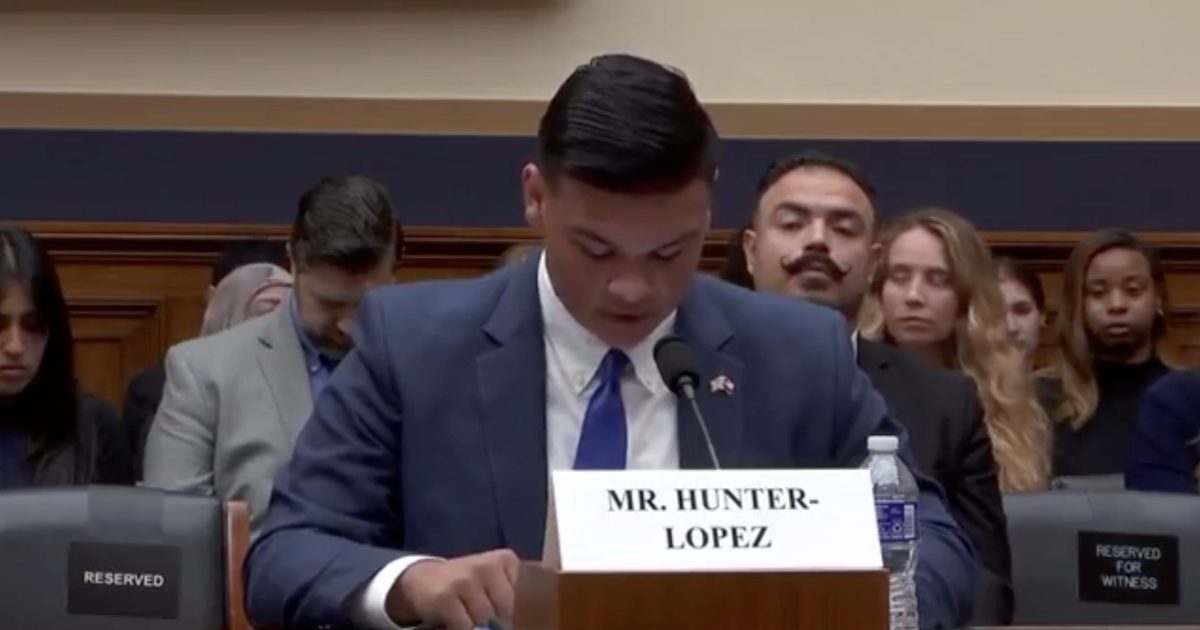 Young man in a suit testifying at a congressional hearing, with audience members engaged in the background.