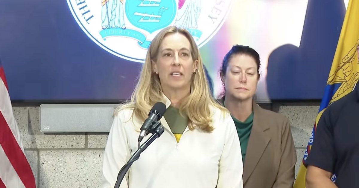 Press conference featuring a female speaker addressing an audience, with state flags and a seal in the background, highlighting key announcements.
