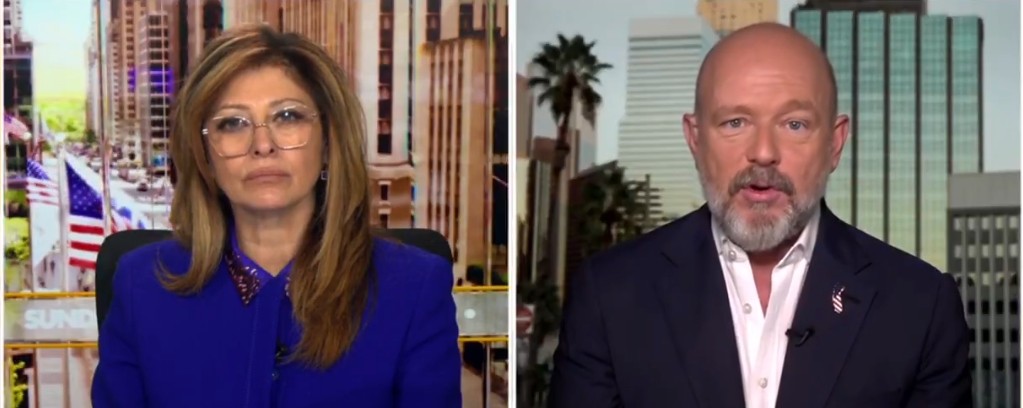 News anchors discussing current events, with a city skyline and American flags in the background, highlighting a professional broadcast setting.
