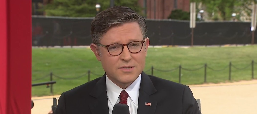 News anchor reporting live outdoors, wearing a suit and glasses, with a blurred background of greenery and barriers.