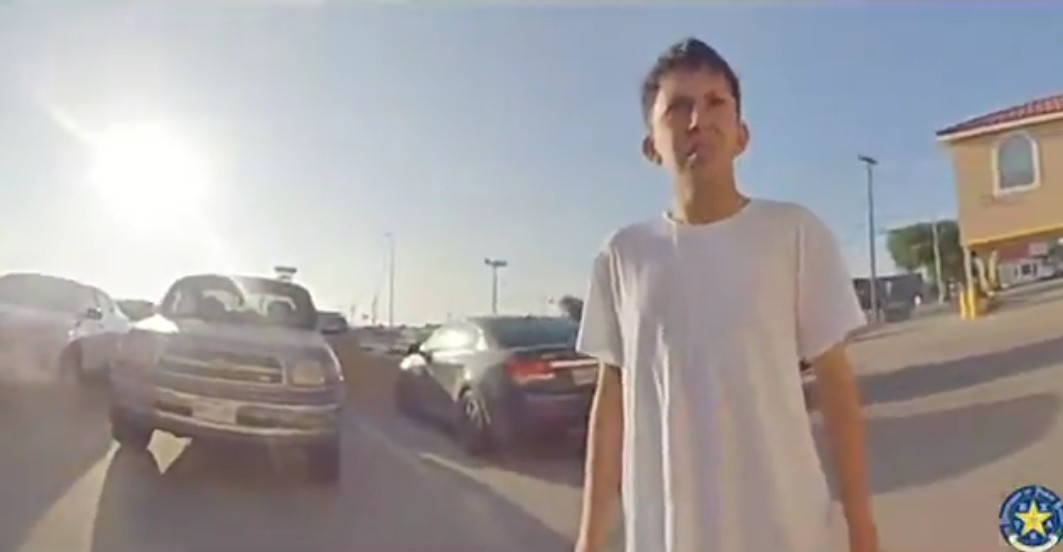 A man in a white t-shirt stands in a parking lot surrounded by vehicles under bright sunlight.