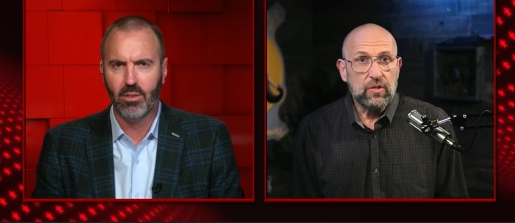 Two men engaged in a serious discussion on a video call, with a red-themed background and a microphone visible in the frame.