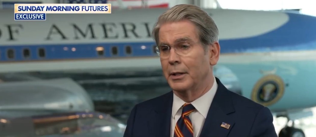 A man in a suit speaks during an interview with an iconic airplane in the background, showcasing the setting of a news segment.