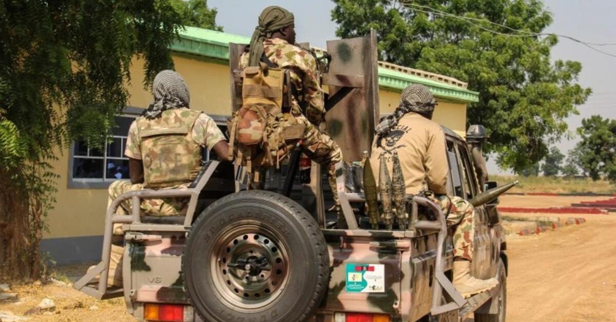 Nigerian soldiers equipped with tactical gear are mounted on a military vehicle, preparing for a mission in a rural area.