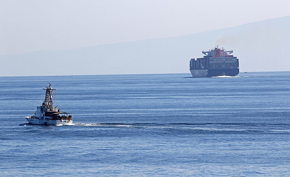 A small boat navigates the calm sea alongside a large cargo ship, highlighting maritime activities in a serene coastal setting.