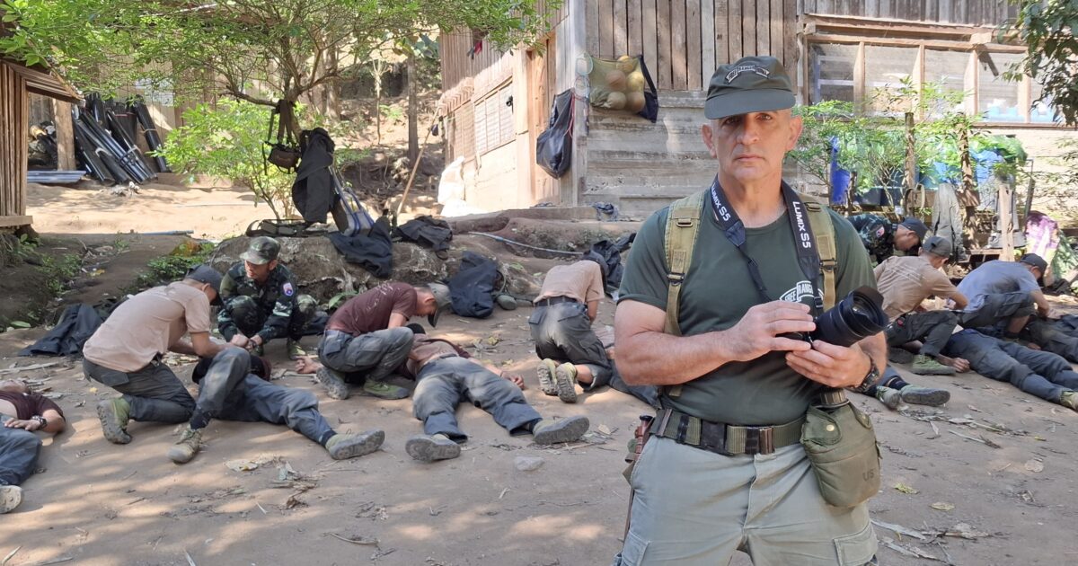 A photographer stands in the foreground while military personnel assist individuals lying on the ground in a wooded area, highlighting a scene of training or rescue operations.