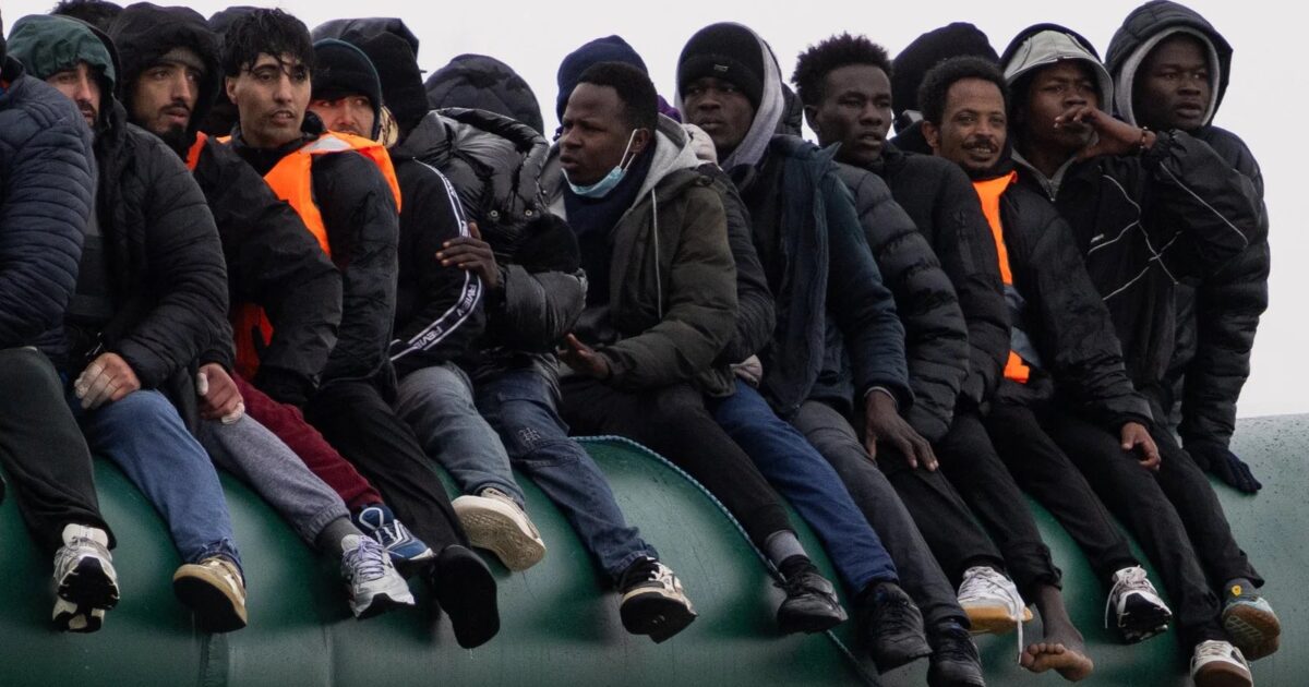 Group of migrants wearing life jackets and winter clothing sitting closely on the edge of a rubber boat in a cold sea.