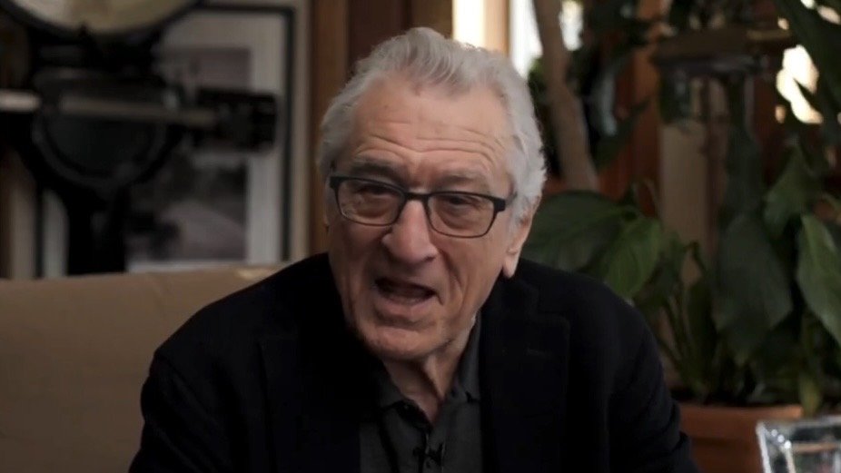Robert De Niro speaks passionately while seated in a cozy setting surrounded by plants and framed photographs.