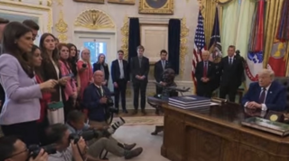Press conference in the Oval Office with Donald Trump seated at the desk, surrounded by reporters and staff, capturing a moment of media interaction.