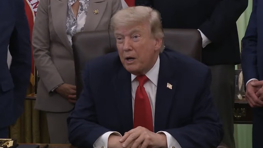 Donald Trump seated at a desk during a formal event, discussing policy with officials and surrounded by American flags.