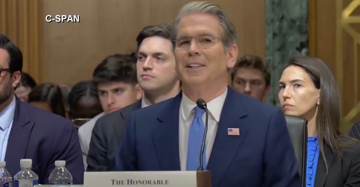 A man in a suit speaks at a Senate hearing, with attentive audience members visible in the background.