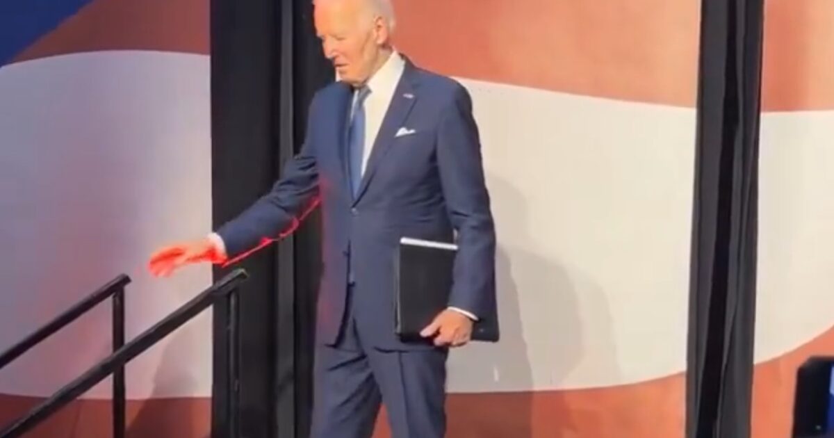 President Biden walking down the stage steps while holding a folder, with a backdrop featuring bold colors.